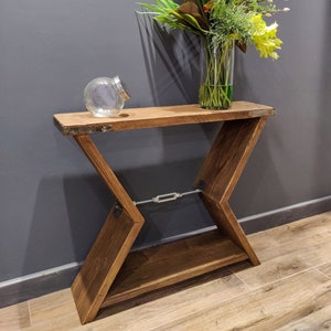 May include: A wooden console table with an X-frame design, stained a warm brown. The table has a lower shelf and is decorated with a clear glass vase filled with yellow and green flowers, and a small glass jar. The table is made of wood.