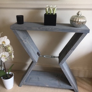 May include: A grey wooden console table with an X-frame design. The table features a rectangular top and a lower shelf. It is decorated with a black candle holder, a black square vase with white flowers, and a silver glass jar.