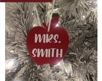 May include: A red apple-shaped Christmas ornament with the text "mrs. SMITH" in white. The ornament hangs from a white ribbon and is displayed on a snow-covered Christmas tree.