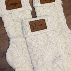 Op de afbeelding: Drie crèmekleurige kerstsokken met kabelgebreid ontwerp. Elke sok heeft een bruin leren label met een naam erop. Er staat "Sargon", "Merry" en "Milano" op.