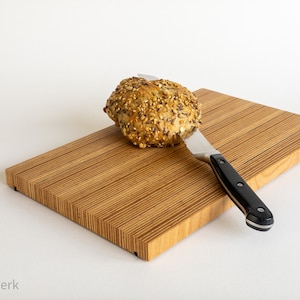 Puede incluir: Una tabla de cortar de madera con un cuchillo y un pan redondo con semillas en la parte superior. La tabla está hecha de finas tiras de madera.