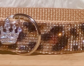 Puede incluir: Un collar de perro de color beige con una hebilla plateada y un dije de corona plateada. El collar está cubierto de diamantes de imitación en un patrón de estampado de leopardo.