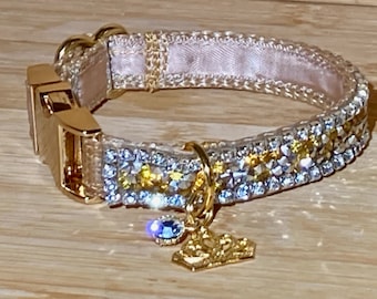 May include: A gold and white dog collar with a crystal and rhinestone design. The collar has a gold buckle and a gold crown charm with a blue crystal.
