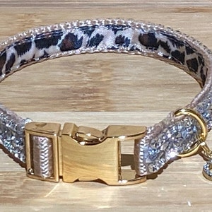 Op de afbeelding: Een glinsterende zilveren hondenhalsband met een gouden gesp en een klein kristallen hartje. De halsband heeft een luipaardprint voering.
