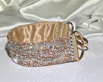 May include: A bedazzled dog collar with a gold-colored metal clasp and rings. The collar is lined with a satin-like fabric and covered in sparkling crystals in shades of gold and silver.