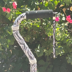 May include: A decorative walking cane with a black, zebra-striped shaft and a black padded handle. The cane is embellished with sparkling rhinestones and a matching wrist strap. Pink flowers and green foliage are in the background.