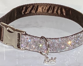 Puede incluir: Un collar de perro marrón con una hebilla plateada y una fila de brillantes de cristal. Un pequeño charm de corona plateada cuelga del collar.