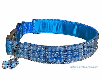 May include: A light blue dog collar with a silver buckle and a charm in the shape of a butterfly. The collar is decorated with rows of white pearls and rhinestones.