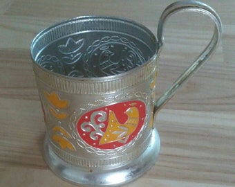 May include: A silver metal cup holder with a red, yellow, and black design. The cup holder has a handle and a decorative pattern.
