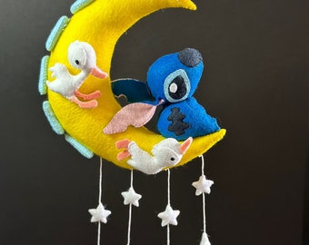 May include: A yellow felt crescent moon with three white felt birds perched on it. A blue felt character with pink ears is hanging from the moon. White felt stars dangle from the bottom of the moon.
