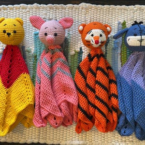 May include: Four handmade crochet baby loveys featuring cartoon character designs. The characters include a yellow bear, a pink pig, an orange tiger, and a blue donkey. Each lovey has a character head and a coordinating blanket in various colors.