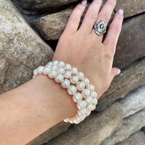 May include: A three-strand pearl bracelet with a cluster of round, off-white pearls. A silver ring with a floral design and a black center stone is also visible. The background is a stone wall.