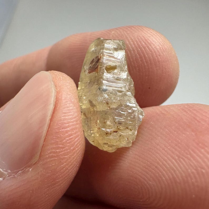 May include: A rough, translucent, light yellow gemstone with a crystalline structure. The gemstone has a slightly irregular shape and appears to be held between two fingers. The stone has a few small, dark inclusions.