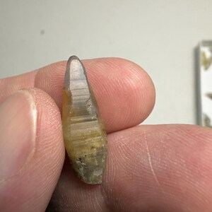 May include: A small, elongated, translucent crystal with a pointed tip and a hexagonal, faceted structure. The mineral displays a gradient of colour, transitioning from a pale, clear top to a warm, golden-yellow base with visible internal inclusions.
