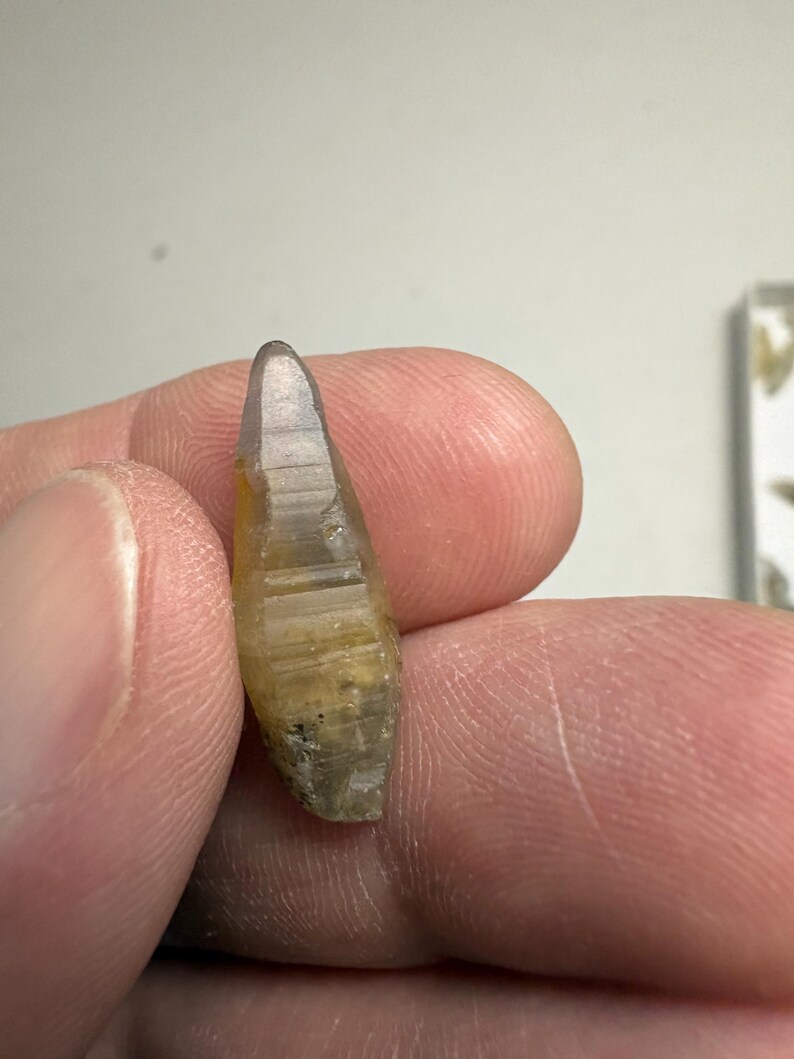 May include: A small, elongated, translucent crystal with a pointed tip and a hexagonal, faceted structure. The mineral displays a gradient of colour, transitioning from a pale, clear top to a warm, golden-yellow base with visible internal inclusions.