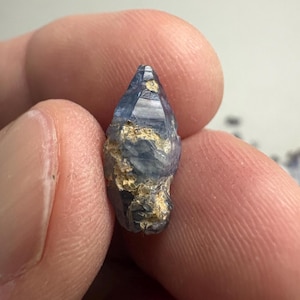 Op de afbeelding: Een ruw, ongeslepen saffierkristal met een puntige, zeshoekige vorm. De steen heeft een diepblauwe kleur met doorschijnende delen en onregelmatige, getextureerde vlekken van lichtbruine of beige minerale insluitingen op het oppervlak.