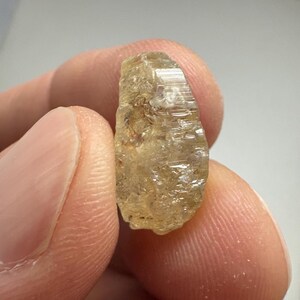 May include: A rough, uncut gemstone with a light yellow-brown hue. The crystal has a textured surface with visible lines and imperfections. The stone is held between two fingers, showcasing its natural, raw form. The overall appearance suggests a mineral specimen.