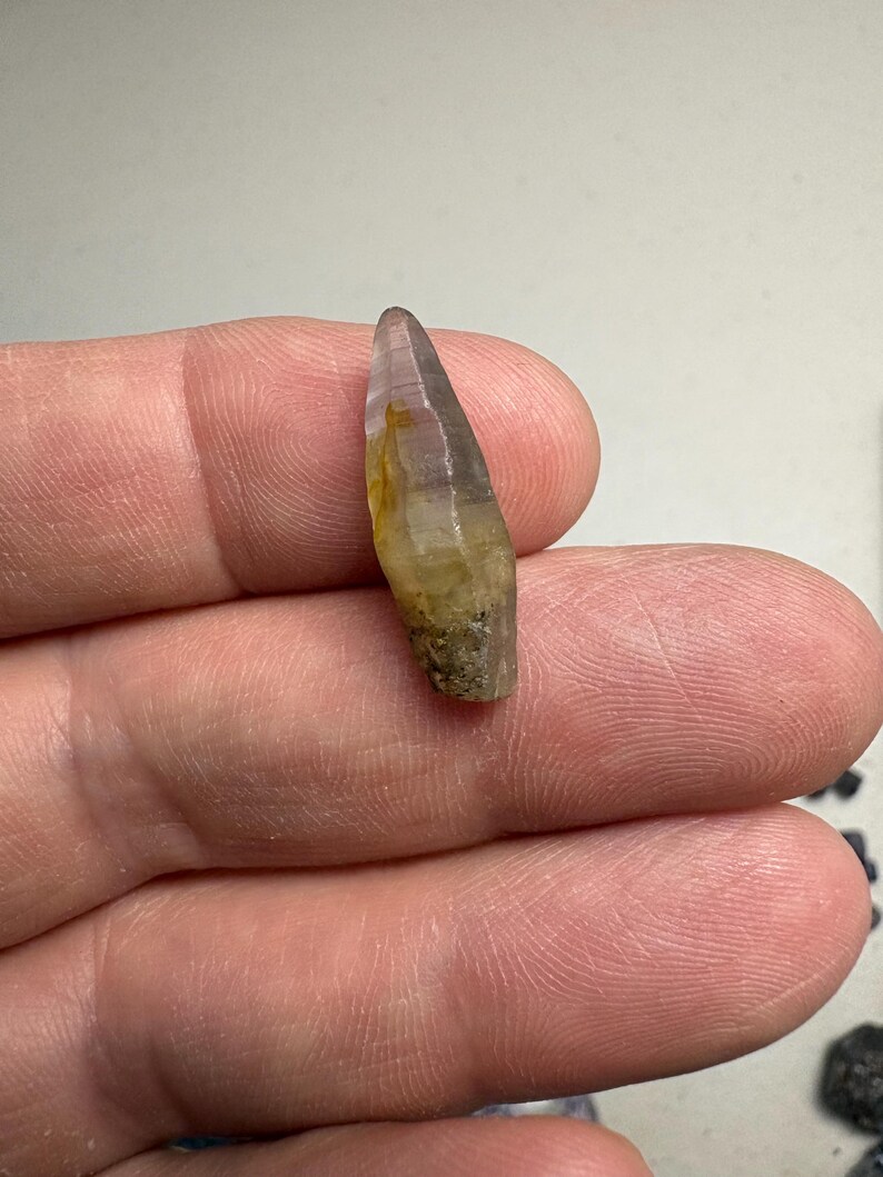 May include: A small, elongated, translucent crystal with a pointed tip. The stone features a colour gradient transitioning from a pale, clear purple at the top to a warm, earthy yellow and brown hue toward the base, which has a rough, textured surface.