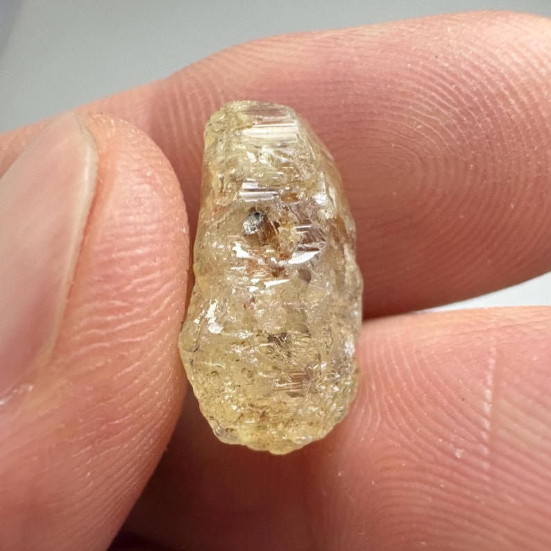 May include: A rough-cut, light yellow gemstone with a crystalline structure. The gem is held between two fingers, showcasing its natural, unpolished appearance. The stone has a slightly translucent quality, with visible internal features.