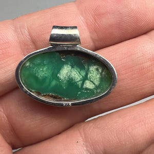 May include: Oval pendant with a vibrant green stone set in a silver bezel. The stone has a unique pattern with lighter green and brown inclusions. The pendant has a silver bail at the top and is marked with "925".