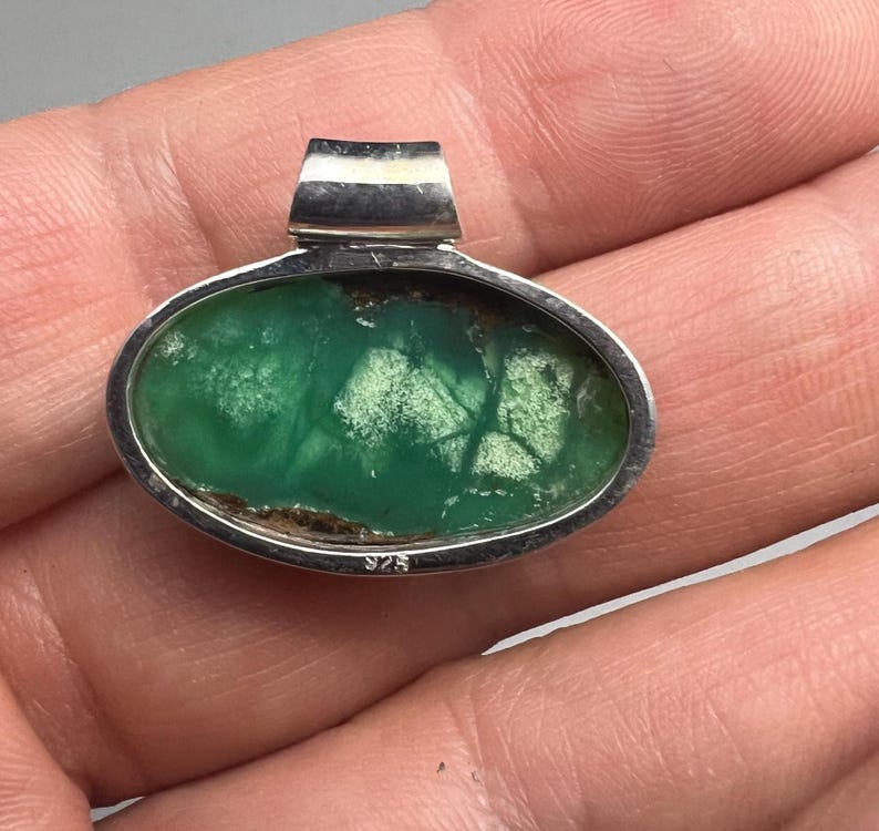 May include: Oval pendant with a vibrant green stone set in a silver bezel. The stone has a unique pattern with lighter green and brown inclusions. The pendant has a silver bail at the top and is marked with "925".