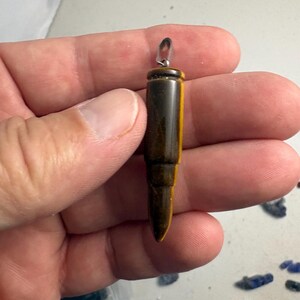 May include: A bullet-shaped pendant carved from brown and gold tiger's eye gemstone. The pendant features a smooth, polished surface with natural banding and a silver-toned metal bail attached to the top for hanging on a chain or cord.