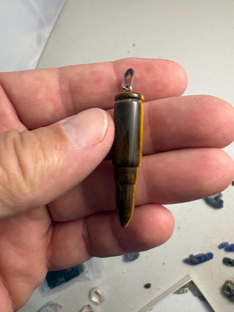 May include: A bullet-shaped pendant carved from brown and gold tiger's eye gemstone. The pendant features a smooth, polished surface with natural banding and a silver-toned metal bail attached to the top for hanging on a chain or cord.