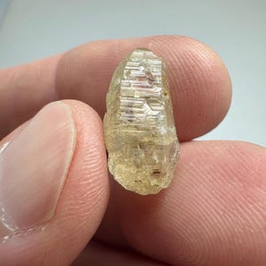 May include: A raw, light yellow-brown crystal with a textured surface. The crystal is held between two fingers, showcasing its natural, unpolished form. The crystal's facets catch the light, revealing its internal structure.