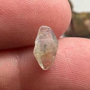 May include: A small, translucent, light-yellow gemstone with a faceted, elongated shape. The gem is held between two fingers, showcasing its clarity and natural form. The stone has subtle internal inclusions.