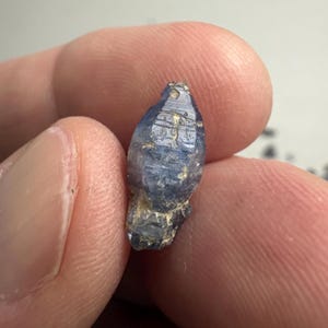 Op de afbeelding: Een kleine, ruwe, langwerpige blauwe saffierkristal met een hexagonale structuur en zichtbare strepen over de lengte. De edelsteen vertoont verschillende tinten diepblauw en doorschijnende gebieden, met een natuurlijke textuur en aardse insluitingen.