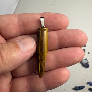 May include: A bullet-shaped pendant carved from golden-brown tiger's eye gemstone, featuring a smooth, polished surface with natural chatoyant banding and a silver-toned metal bail attached to the top for hanging on a necklace chain.