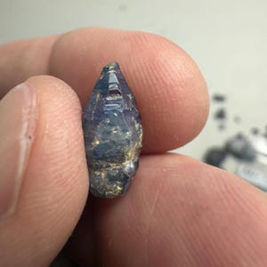 Op de afbeelding: Een kleine, ruwe, langwerpige saffierkristal met een puntige top en een hexagonale structuur. De edelsteen vertoont een diepblauwe kleur met doorschijnende delen en natuurlijke, ruwe, getextureerde oppervlakken langs de basis en zijkanten.