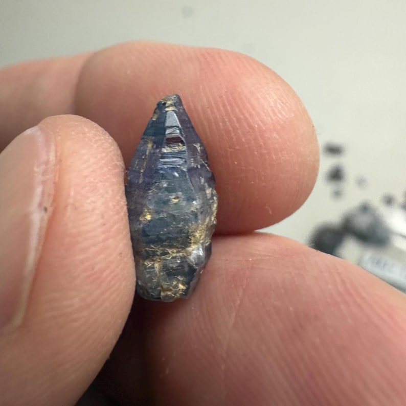 Op de afbeelding: Een kleine, ruwe, langwerpige saffierkristal met een puntige top en een hexagonale structuur. De edelsteen vertoont een diepblauwe kleur met doorschijnende delen en natuurlijke, ruwe, getextureerde oppervlakken langs de basis en zijkanten.