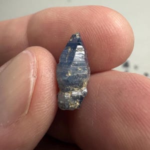 Op de afbeelding: Een kleine, ruwe, langwerpige saffierkristal met een diepblauwe kleur en zichtbare natuurlijke strepen langs de verticale facetten. De steen heeft een puntige bovenkant en een licht onregelmatige, getextureerde basis met lichte minerale insluitingen.