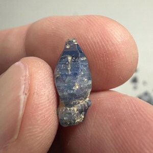 Op de afbeelding: Een kleine, ruwe saffierkristal met een diepblauwe kleur en een natuurlijk gefacetteerde, langwerpige vorm. De steen heeft zichtbare horizontale strepen op het oppervlak, met vlekken van lichter blauw en aardse, beige minerale insluitingen.
