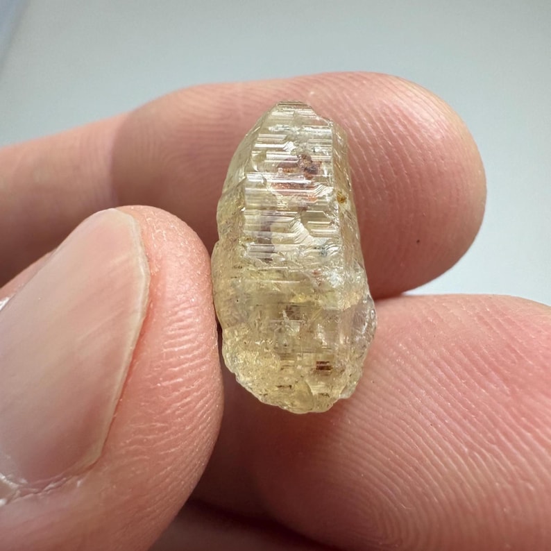 May include: A translucent, light yellow crystal with a rough, textured surface. The crystal has a rectangular shape with visible internal structures and a slightly rounded base. Small reddish-brown inclusions are scattered throughout.