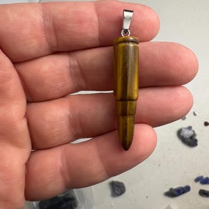 May include: A bullet-shaped pendant carved from golden-brown tiger's eye gemstone, featuring a polished, chatoyant surface with dark brown banding and a silver-toned metal bail attached to the top for hanging on a chain or cord.