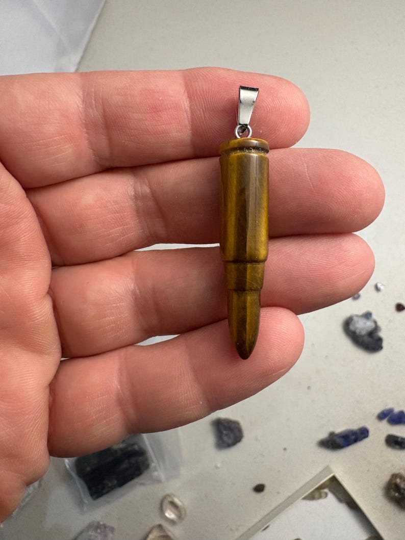 May include: A bullet-shaped pendant carved from golden-brown tiger's eye gemstone, featuring a polished, chatoyant surface with dark brown banding and a silver-toned metal bail attached to the top for hanging on a chain or cord.
