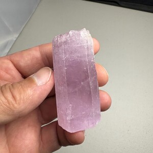 K&ouml;nnte beinhalten: Ein hellvioletter Kristall mit rechteckiger Form. Der Kristall hat eine raue, strukturierte Oberfl&auml;che und wird in einer Hand gehalten. Der Kristall hat eine weiche, pastellviolette Farbe.