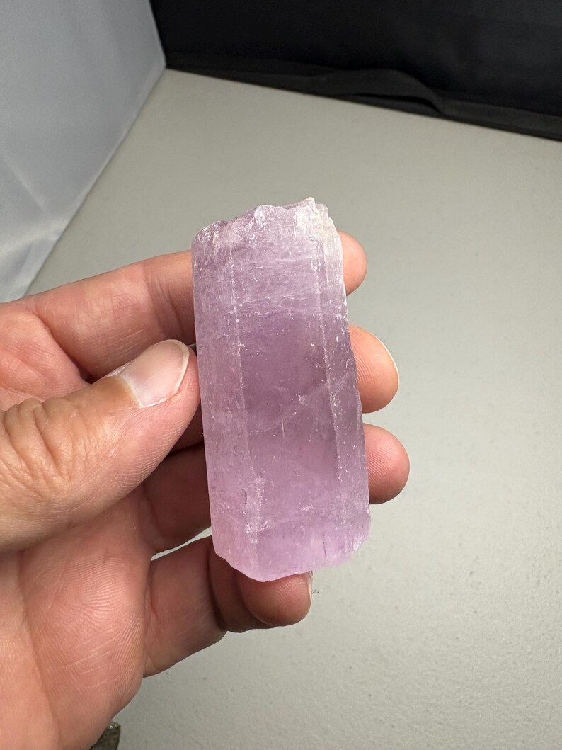 K&ouml;nnte beinhalten: Ein hellvioletter Kristall mit rechteckiger Form. Der Kristall hat eine raue, strukturierte Oberfl&auml;che und wird in einer Hand gehalten. Der Kristall hat eine weiche, pastellviolette Farbe.
