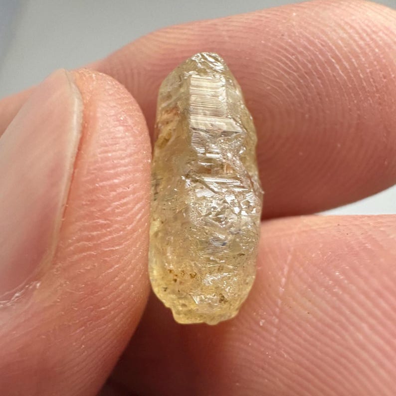 May include: A rough, light-yellow crystal with a textured surface. The crystal is held between two fingers, showcasing its natural, unpolished form. The gemstone has a translucent quality, catching the light.