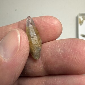 May include: A small, elongated, double-terminated crystal with a translucent appearance. The stone features a gradient of colours, transitioning from a pale, clear purple at the pointed tip to a warm, earthy yellow and brown hue toward the base.