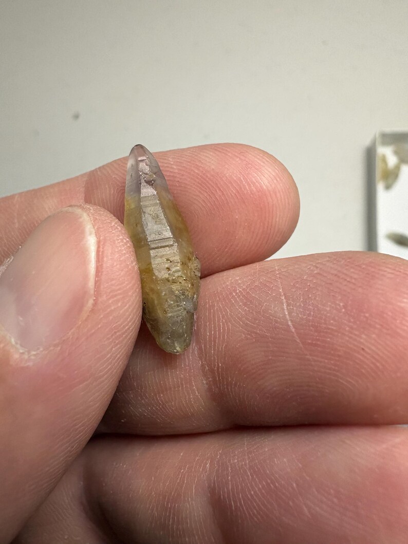 May include: A small, elongated, double-terminated crystal with a translucent appearance. The stone features a gradient of colours, transitioning from a pale, clear purple at the pointed tip to a warm, earthy yellow and brown hue toward the base.