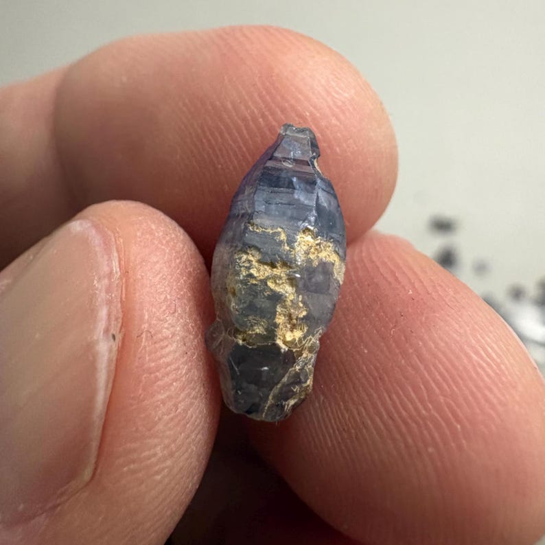 Op de afbeelding: Een kleine, langwerpige, doorschijnende blauwe saffierkristal met een hexagonale structuur en zichtbare natuurlijke insluitingen. De ruwe edelsteen heeft een gefacetteerde, taps toelopende punt aan de bovenkant en een ruw oppervlak met lichtbruine mineraalafzettingen.