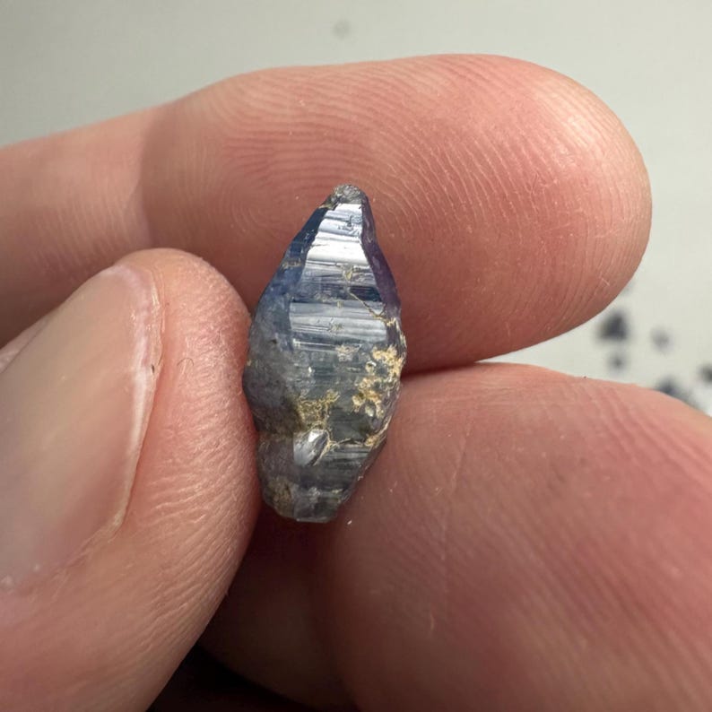 Op de afbeelding: Een kleine, ruwe, langwerpige saffierkristal met een diepblauwe tint en zichtbare natuurlijke strepen langs de verticale facetten. De steen heeft een puntige bovenkant en een onregelmatige, getextureerde basis met lichte minerale insluitingen.