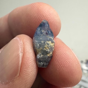 Op de afbeelding: Een kleine, langwerpige, ruwe saffierkristal met een zeshoekige, taps toelopende vorm. De steen heeft een doorschijnende blauwe kleur met zichtbare horizontale strepen over de lengte en vlekken van lichtbruine, aardse mineraalinsluitingen.