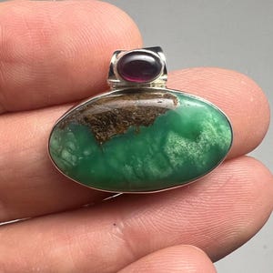 May include: Oval pendant with a vibrant green stone, accented by a smaller, dark red stone at the top. The pendant has a silver-colored metal setting. The green stone has brown inclusions.