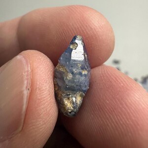 Op de afbeelding: Een kleine, ruwe, langwerpige blauwe saffierkristal met een hexagonale structuur. De steen vertoont een mix van doorschijnende blauwe secties en ondoorzichtige, aardebruine minerale insluitingen op het oppervlak, met natuurlijke facetten.