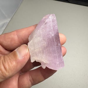 K&ouml;nnte beinhalten: Eine Hand h&auml;lt ein rohes, kristallines Mineralexemplar. Der Stein hat eine blasslavendelfarbene Farbe mit einer durchscheinenden Qualit&auml;t. Der Kristall hat eine raue, nat&uuml;rliche Textur und ein geschichtetes Aussehen.
