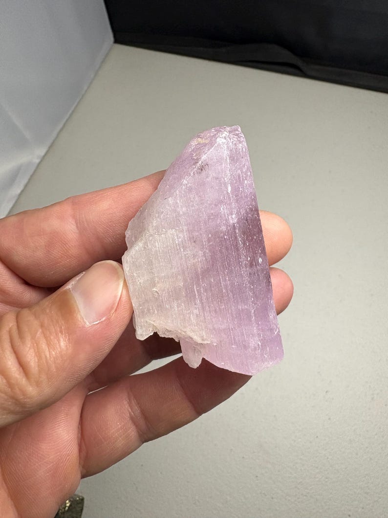 K&ouml;nnte beinhalten: Eine Hand h&auml;lt ein rohes, kristallines Mineralexemplar. Der Stein hat eine blasslavendelfarbene Farbe mit einer durchscheinenden Qualit&auml;t. Der Kristall hat eine raue, nat&uuml;rliche Textur und ein geschichtetes Aussehen.